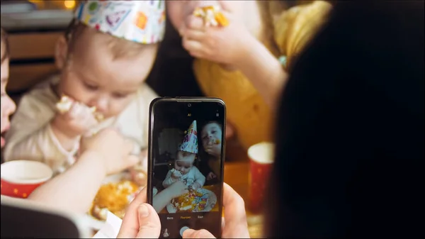 One-year-old girls birthday party. Mom brings a plate of cake. The sisters happily eat the cake and get dirty with the cake. My mother is filming the holiday on her phone.