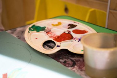 Sanat eserleriyle çizim sınıfı. 4-8 yaş arası çocukların olduğu bir sınıf. Bir kız yeşil bir kağıda parlak renkli bir resim çiziyor.