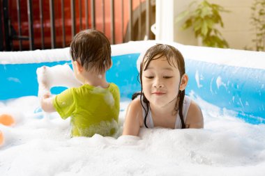 Kız ve erkek kardeş evdeki yüzme havuzunda baloncuklarla oynarken eğleniyorlar. Yaz su oyunu, aile mutluluğu, çocukların mutluluğu.