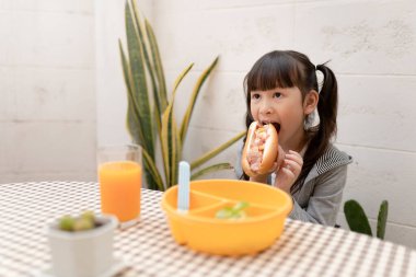 Genç Asyalı kız kahvaltısını ve portakal suyunu evde hala mutlu bir şekilde yiyor. Sağlıklı beslenme, yemek ve atıştırma, jambonlu ve peynirli sandviç, çocuk mutlu beslenme