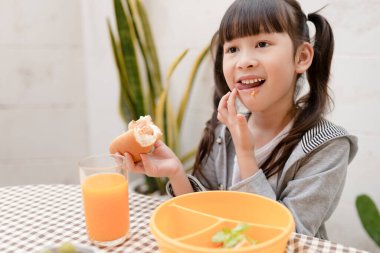 Genç Asyalı kız kahvaltısını ve portakal suyunu evde hala mutlu bir şekilde yiyor. Sağlıklı beslenme, yemek ve atıştırma, jambonlu ve peynirli sandviç, çocuk mutlu beslenme