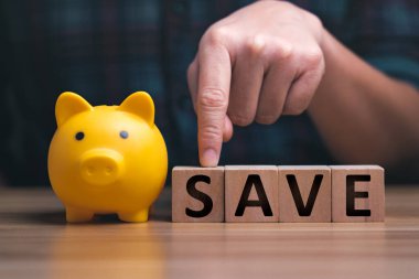 Para biriktiriyorum. Adamın eli ahşap bir masa üzerindeki tahtadan bir bloktan tasarruf edilen paraya, servetten tasarruf edilen paraya ve finansal konseptlere iş finansmanına finansal planlamaya işaret ediyor.