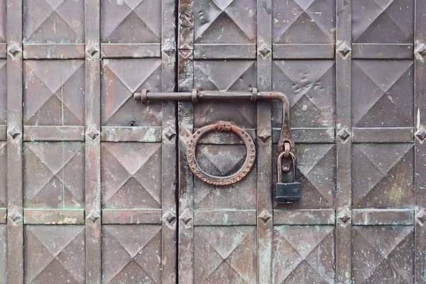An old wrought iron door knocker, a metal latch and a lock on an old metal door in the Russian style.  The forged hammer handle and the handmade latch are made of iron in close-up against the background of a metal door.