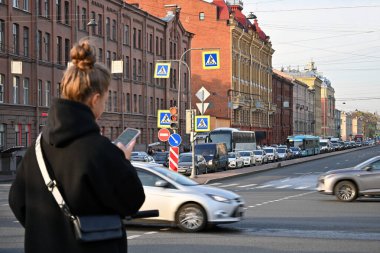 Siyah ceketli genç bir kız şehir merkezinde arabalı bir caddenin arka planında telefonuna bakıyor..     