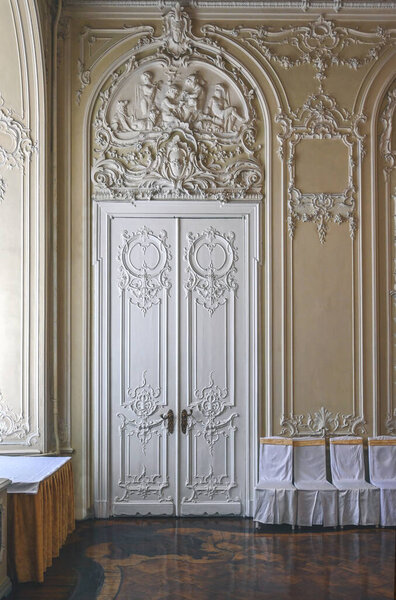 An old wooden white door with carvings, with a decorative stucco panel with sculptures and a cartouche, in a restored historical architectural interior with richly decorated walls in Rococo, Baroque, eclectic style. 