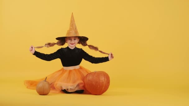 Kleines blondes Mädchen im Hexenkostüm an Halloween vor orangefarbenem Studiohintergrund