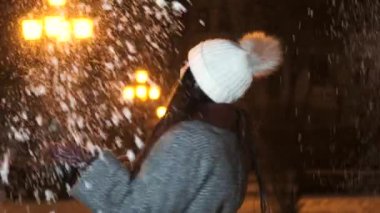 Kış kıyafetleri içinde mutlu genç bir kadın akşam kentinde kar yağdırıyor.