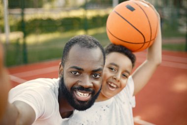 Siyahi baba ve çok ırklı oğlu basketbol sahasında durup selfie çekiyorlar.