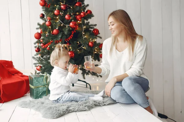 Madre con hija pequeña sentada cerca del árbol de Navidad y bebiendo jugo. — Foto de Stock