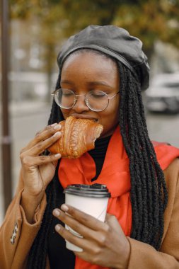 Paltolu genç siyah kadın dışarıda kruvasanla kahve içiyor.