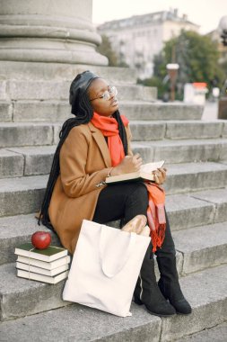 Paltolu genç siyah kadın merdivenlerde oturuyor ve kitap okuyor.