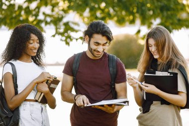 Three international students standing in a park and holding a books