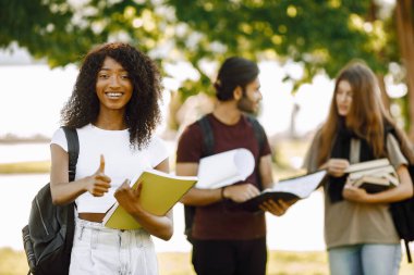 Three international students standing in a park and holding a books