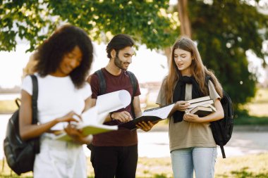 Three international students standing in a park and holding a books