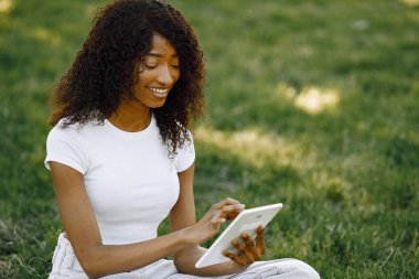 Female african student sitting on a grass and using a tablet
