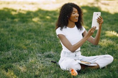 Female african student sitting on a grass and using a tablet fo a video call