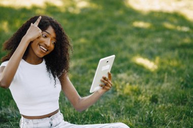 Female african student sitting on a grass and using a tablet fo a video call