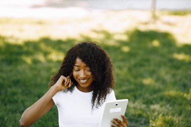 Female african student sitting on a grass and using a tablet fo a video call