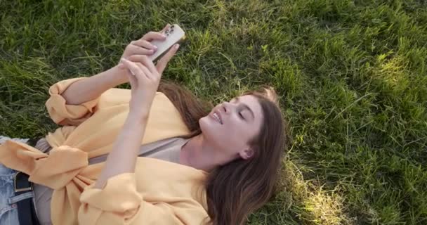 Younng woman with smartphone lying on grass — стоковое видео