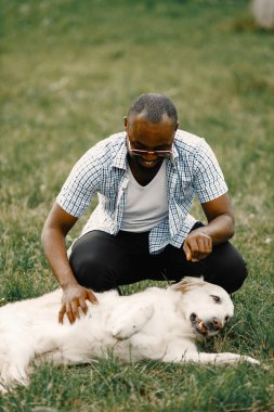 Güneş gözlüklü siyahi bir adamın köpeği çimlerin üzerinde oturuyor.