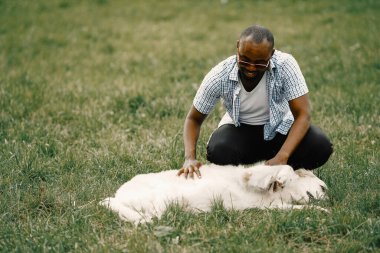 Güneş gözlüklü siyahi bir adamın köpeği çimlerin üzerinde oturuyor.