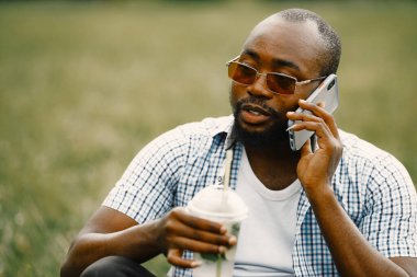 Güneş gözlüklü siyah Amerikalı bir adam çimlerin üzerinde oturmuş telefonla konuşuyor.