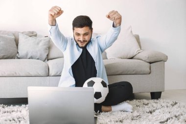 Esmer adam yerde oturuyor ve dizüstü bilgisayarında futbol izliyor.