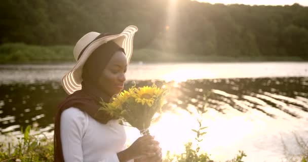 Svart kvinna i hatt sitter på filt vid floden — Stockvideo