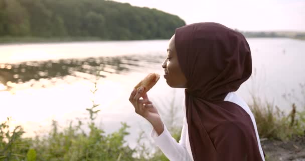 Müslüman siyah kadın nehir kıyısında kruvasan yiyor. — Stok video