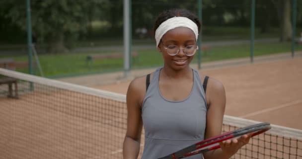 Zwarte vrouw in bril staan met raket op tennisbaan — Stockvideo