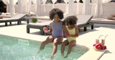 İki küçük kız havuz kenarında oturup karpuz yiyorlar.