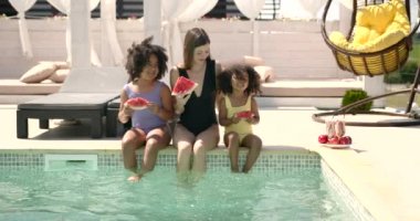 İki küçük kız havuz kenarında oturup karpuz yiyorlar.