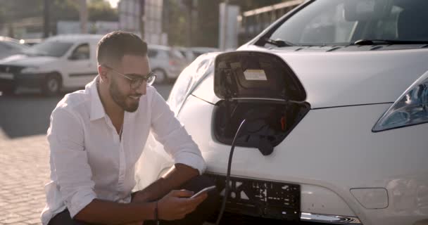 Mladý muž používající mobilní telefon při nabíjení elektrického auta — Stock video