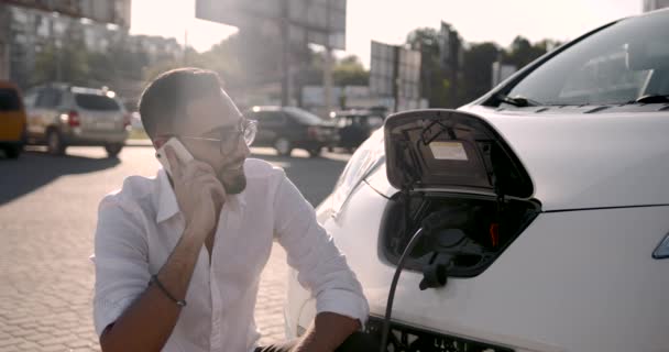 Jeune homme utilisant un téléphone portable tout en rechargeant une voiture électrique — Video