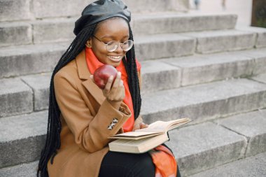 Elinde kitap ve elmayla merdivenlerde oturan montlu siyah bir kadın.