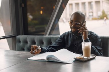 Bir kafede elinde bir fincan kahveyle oturan siyah bir adam ve bir telefon kullanıyor.