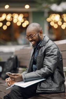 Siyah bir adam bankta oturuyor ve telefon kullanıyor.