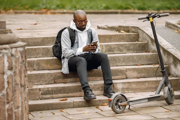 Uomo nero utilizzando un telefono mentre seduto su una strada con uno scooter — Foto Stock