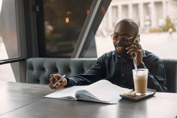 Homem preto sentado em um café com uma xícara de latte e usando um telefone — Fotografia de Stock