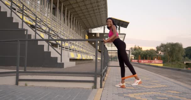 Mujer negra motivada feliz haciendo ejercicios para las piernas con banda de goma — Vídeos de Stock