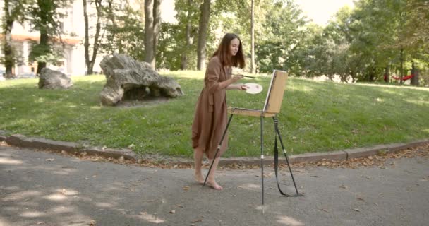 Konstnären står på huk framför ett staffli i parken — Stockvideo