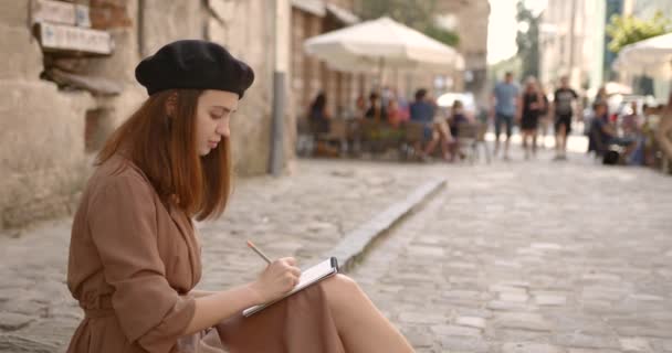 Девушка-художник сидит на тротуаре и рисует в блокноте — стоковое видео