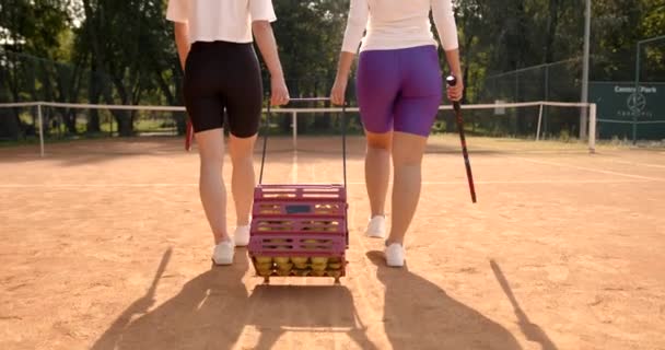 Dos mujeres llevando caja para pelotas de tenis a lo largo de la cancha — Vídeo de stock