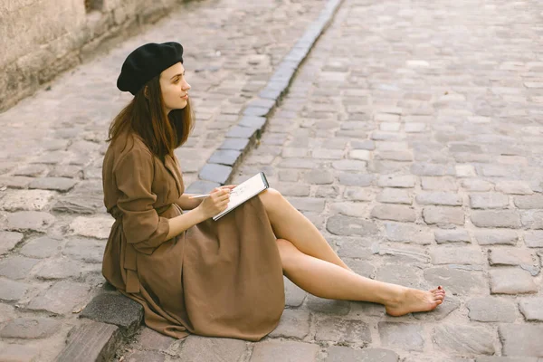 Dívka v šatech a černý klobouk malování náčrtek, zatímco sedí na ulici — Stock fotografie
