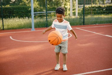 Basketbol sahasında dikilip turuncu bir topla oynayan çok ırklı bir çocuk.