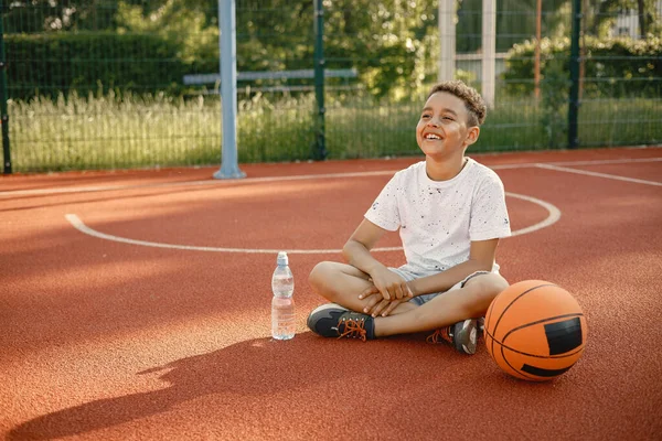 Rapaz multirracial sentado num campo de basquetebol com uma garrafa de água — Fotografia de Stock