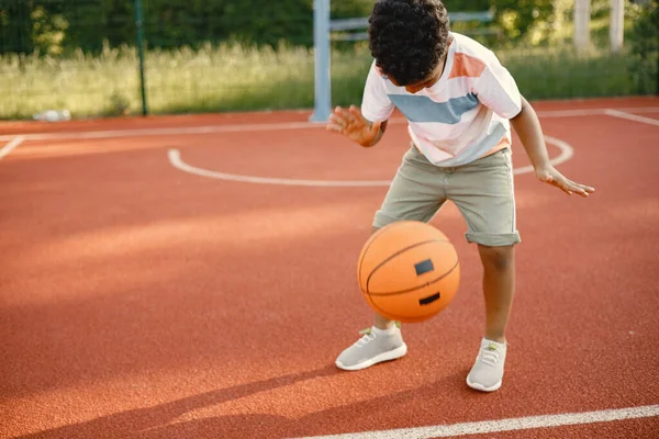 Rapaz multirracial de pé em um campo de basquete e jogar com uma bola laranja — Fotografia de Stock