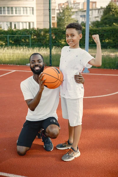 Siyahi baba ve çok ırklı oğlu basketbol sahasında yan yana duruyorlar.
