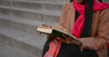 Genç siyah bir kız sokakta merdivenlerde oturuyor ve kitap okuyor..