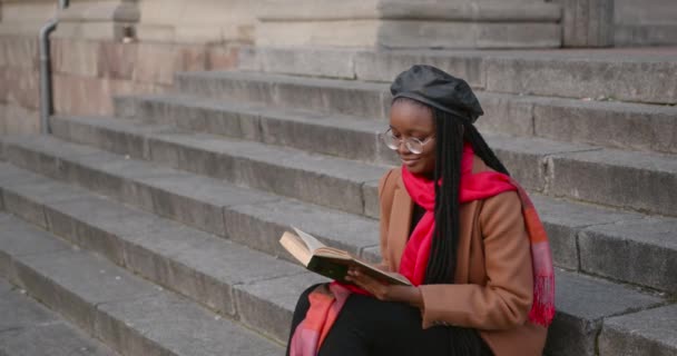 Молода чорна дівчина сидить на сходах на вулиці і читає книгу . — стокове відео